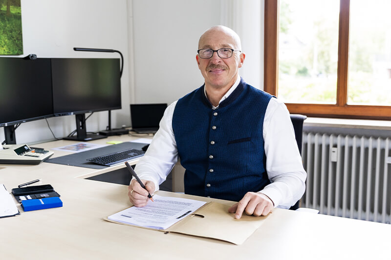 Herr Göbel sitzt an einem Schreibtisch. Er hält einen Stift und hat ein Dokument vor sich