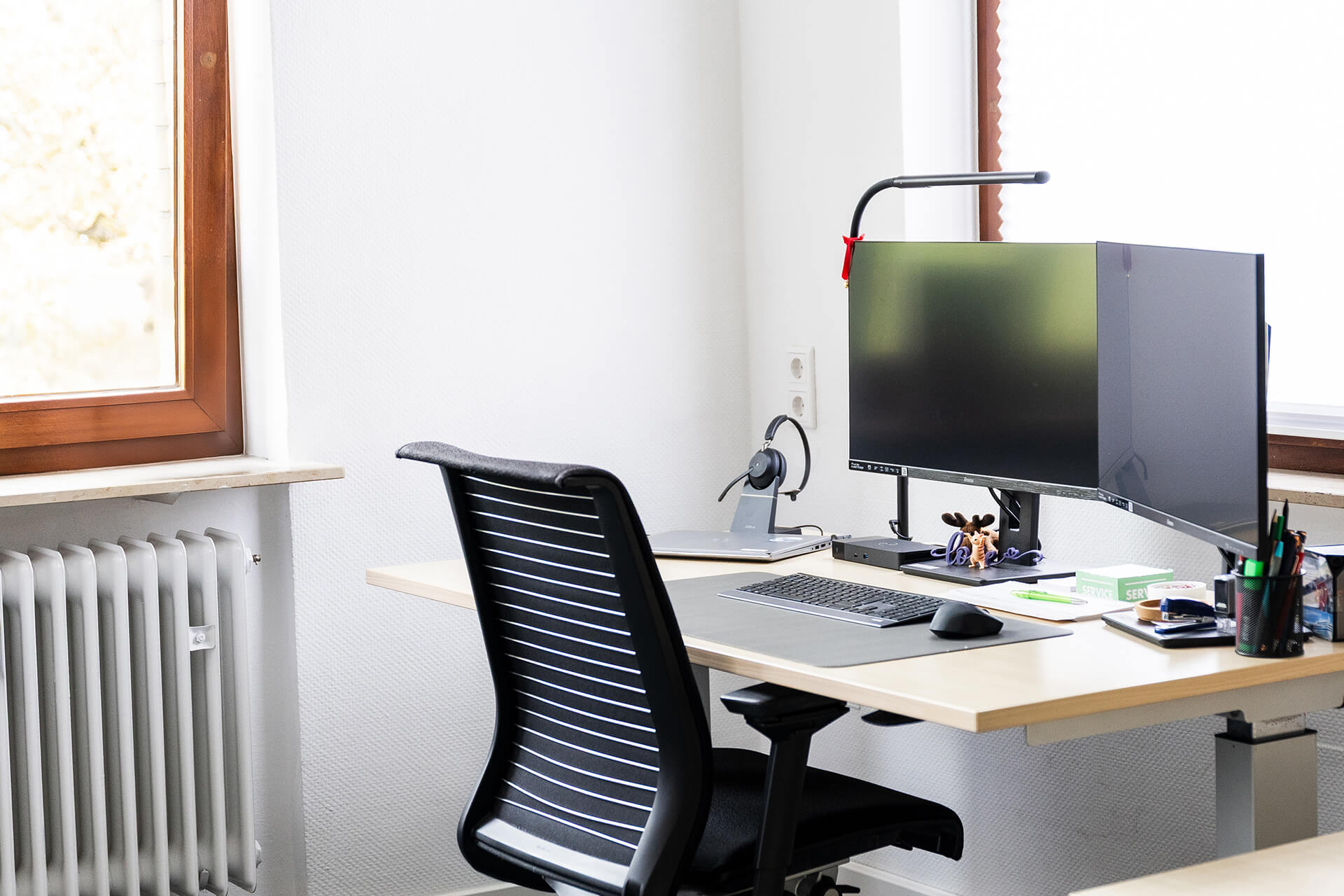 Ein Schreibtisch mit einem schwarzen Bürostuhl, einem Monitor, einer Schreibtischlampe und verschiedenen Büromaterialien. Ein Fenster mit Holzrahmen ist im Hintergrund.