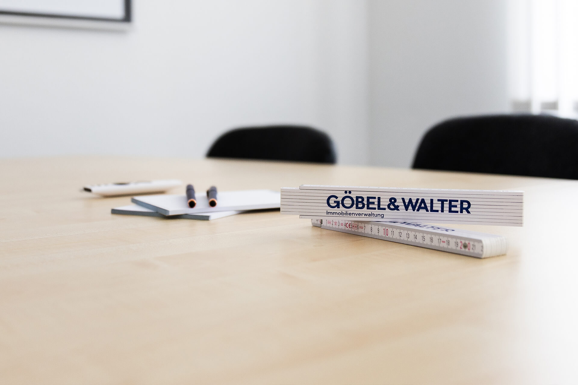 Auf einem Tisch liegen zwei Bleistifte, ein Notizblock und zwei Lineale mit der Aufschrift "GÖBEL & WALTER". Im Hintergrund sind zwei Stühle sichtbar.